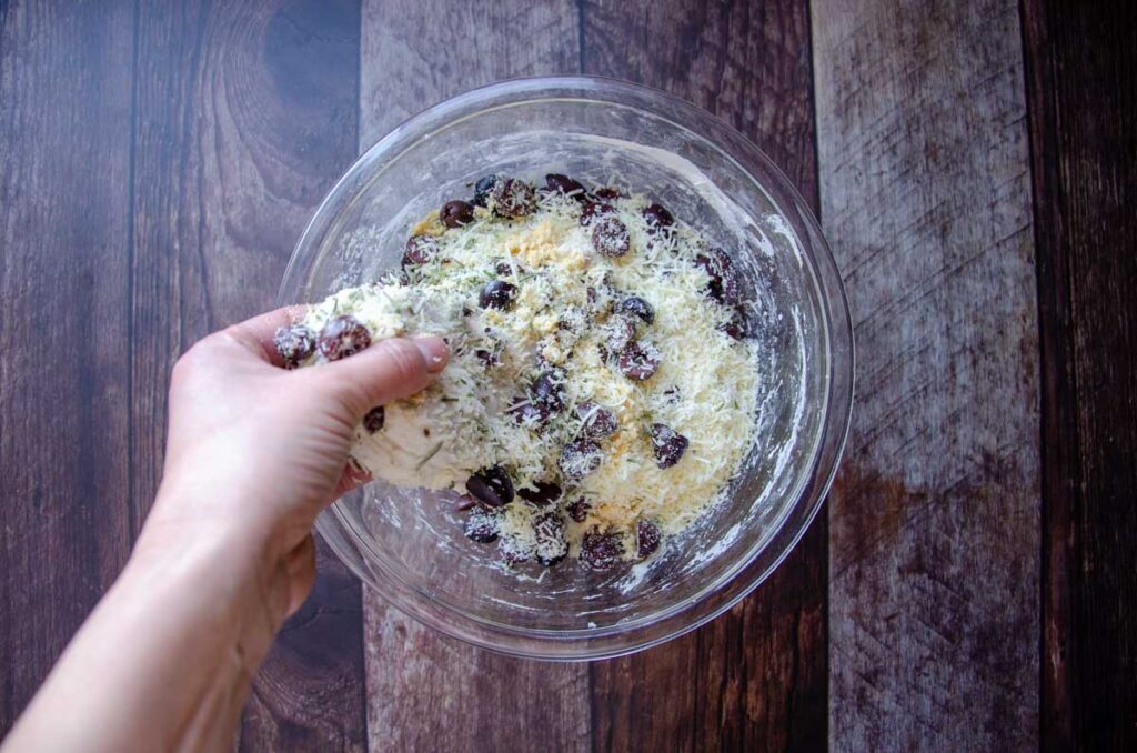 adding olives, cheese, and herbs into sourdough bread dough in a glass bowl