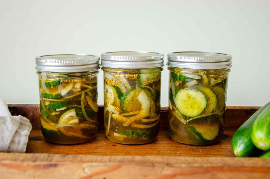 jars of bread and butter pickles next to some whole cucumbers