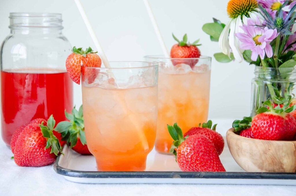 two glasses of strawberry shrubs, flowers, and a jar of straberry shrub mixture