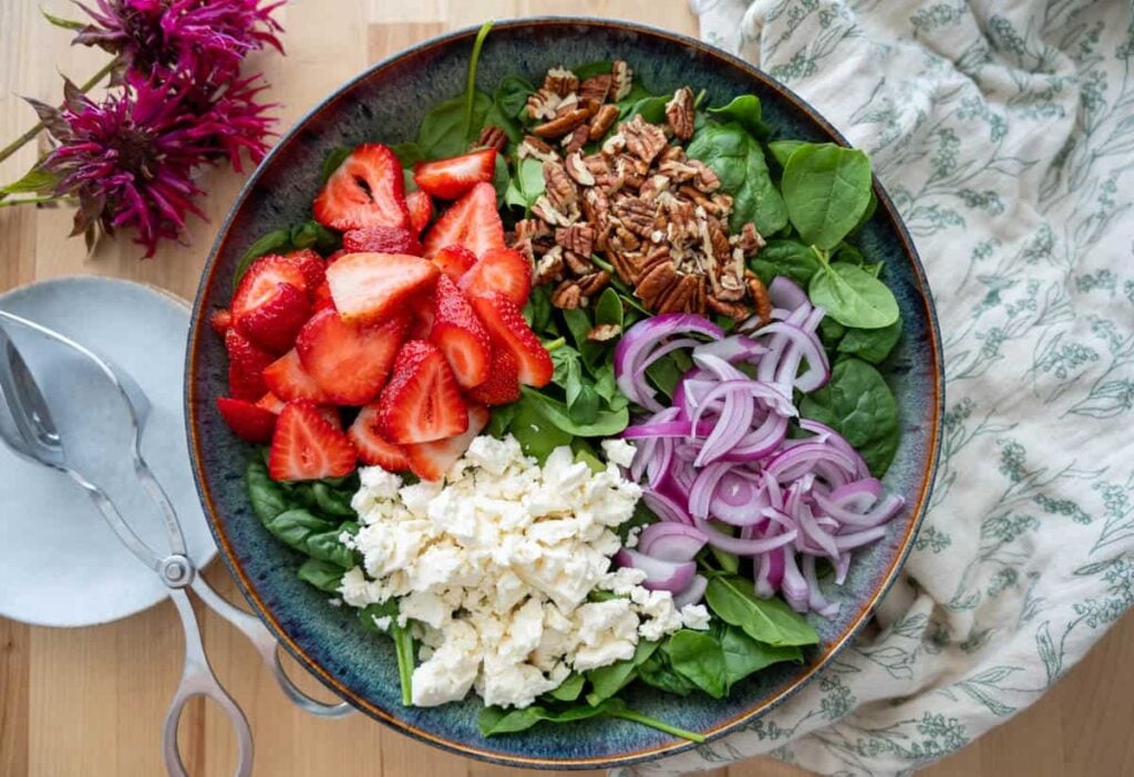 a bed of spinach topped with strawberries, pecans, red onion, and crumbled feta cheese in a large salad bowl
